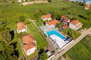 An aerial view of a property with multiple buildings, a swimming pool, and a large green field. - Kamari Corfu Hotel & Apartments (Corfu)
