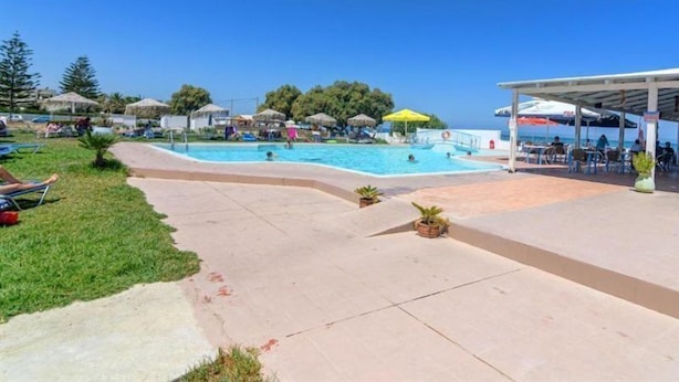 A pool area with lounge chairs and umbrellas, surrounded by a covered seating area and a building with a thatched roof. - Varvaras Diamond (Rethymno)
