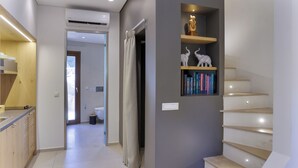 A modern interior with a staircase, a bookshelf with decorative items, and a kitchen area. - Edem Garden Residence (Mykonos)