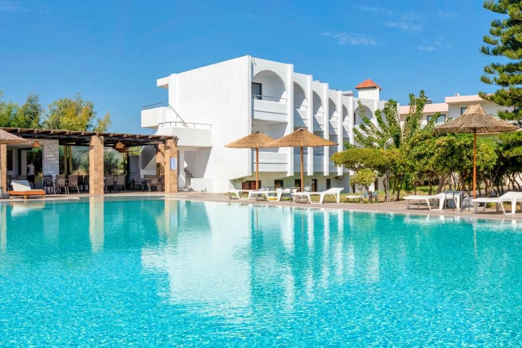 A clear blue pool with lounge chairs and umbrellas, surrounded by a white building with balconies.