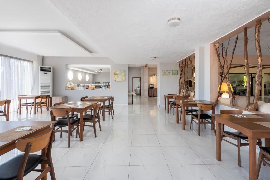A spacious dining area with wooden tables and chairs, a white tiled floor, and a view of the kitchen in the background.