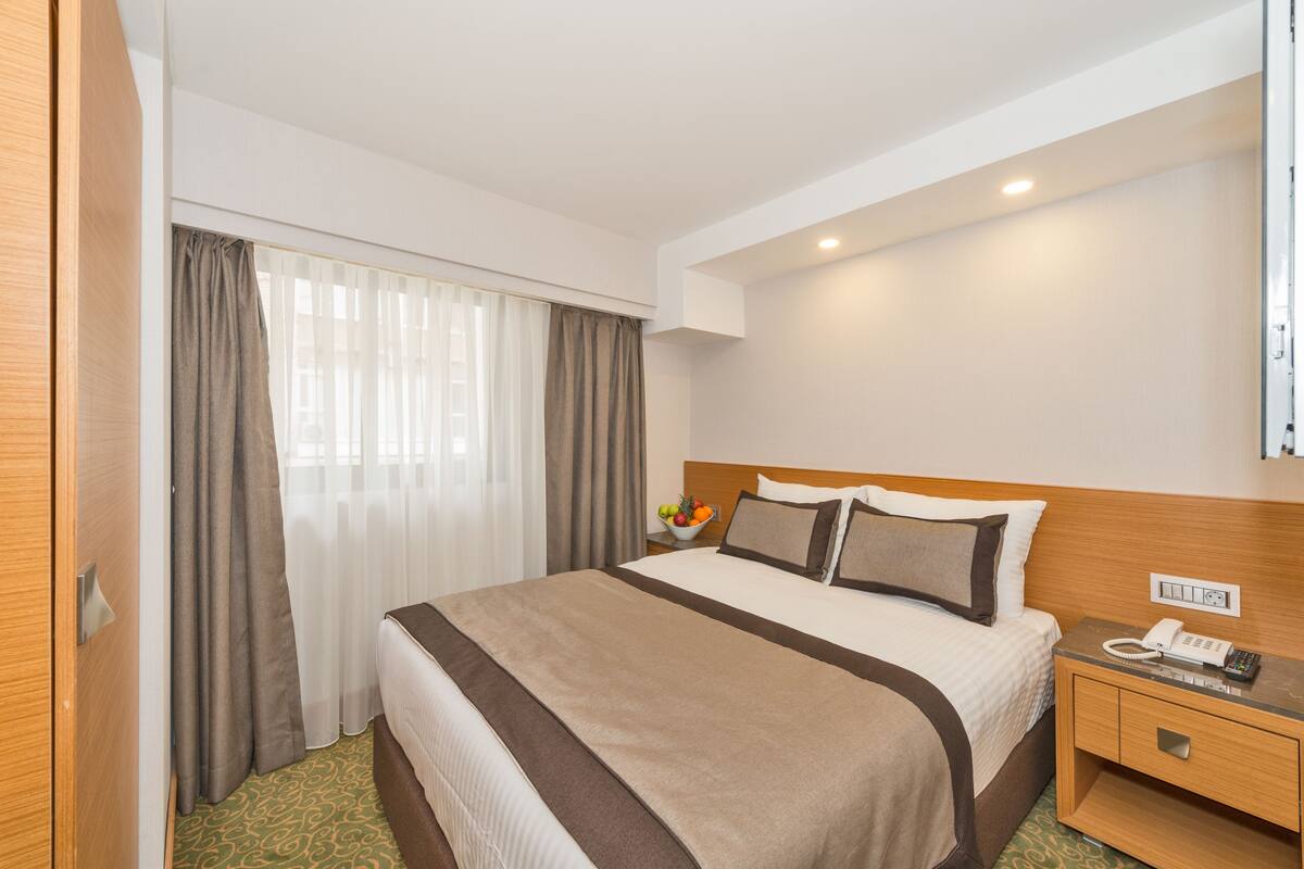 A hotel room with a large bed, a bedside table with a phone, a bowl of fruit, and a window with curtains.