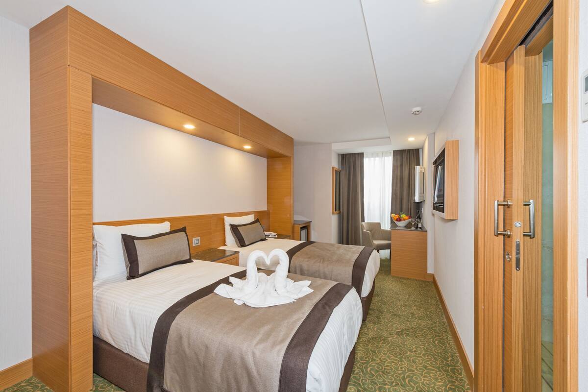 A hotel room with two beds, wooden headboards, a flat-screen TV, a desk with a chair, and a bathroom visible through a glass door.