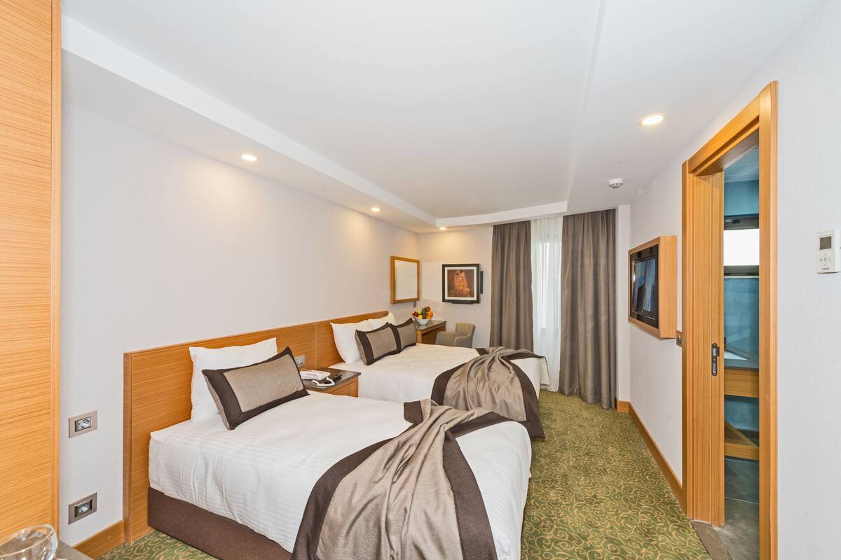 A hotel room with two beds, a desk, a chair, a small table with a vase, and a window with curtains.