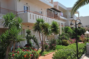 Un edificio residencial con varios balcones, un jardín con diversas plantas y flores, y un camino que lleva a la entrada.
