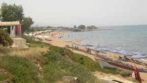A beach with rows of sunbeds and umbrellas, a small building with a red roof, and boats along the shore. - Oasis Scaleta Hotel (Rethymno)