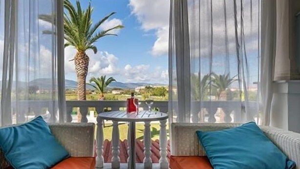 A balcony with two cushioned seating areas, a small table, and a view of palm trees and mountains. - Hotel Sirocco - Adults Only (Zakynthos)