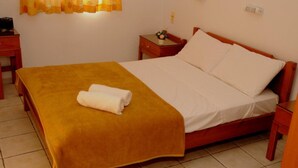 A neatly made bed with a yellow blanket and white pillows, a small bedside table with a lamp, and a window with yellow curtains. - Matheos Apartments (Hersonissos)