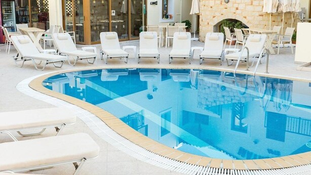 A pool area with lounge chairs and umbrellas, a stone wall, and a Coca-Cola refrigerator. - Matheos Apartments (Hersonissos)