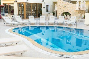 A pool area with lounge chairs and umbrellas, a stone wall, and a Coca-Cola refrigerator. - Matheos Apartments (Hersonissos)