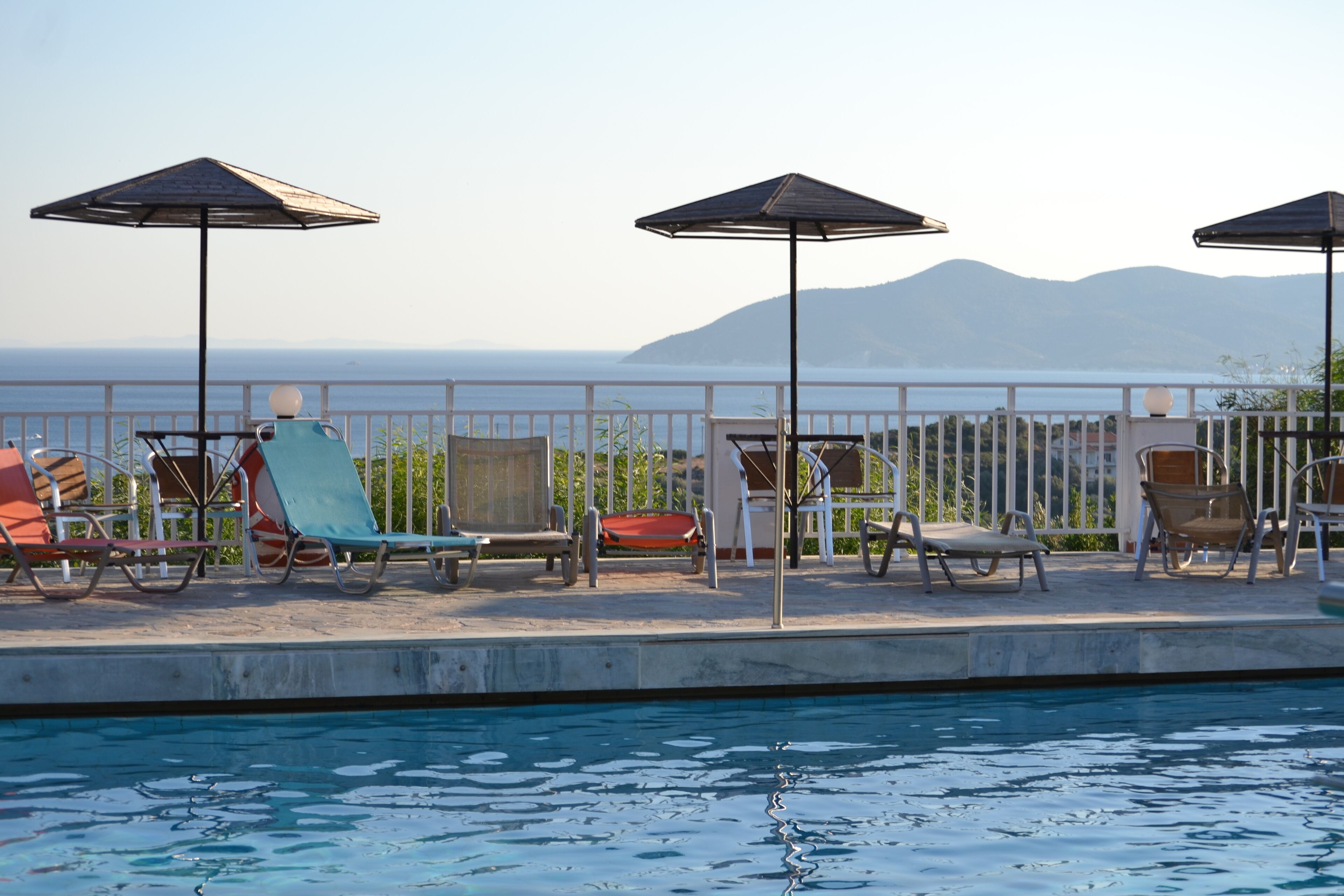 A poolside area with lounge chairs and umbrellas, a clear view of the sea, and a mountainous backdrop.