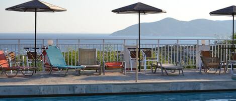 A poolside area with lounge chairs and umbrellas, a clear view of the sea, and a mountainous backdrop.