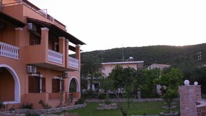A multi-story building with balconies, a garden with stone planters, and a hilly landscape in the background. - Vasso Studios (Corfu)