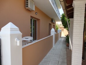 A narrow outdoor corridor with a white railing, a small table with a metal bowl, and a door leading to a balcony. - Makis Louvlis Luxury Apartments (Corfu)