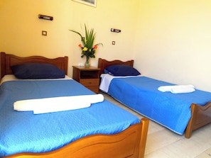 A hotel room with two single beds, each with a blue bedspread and white towels. There is a wooden nightstand with a vase of flowers between the beds. - Makis Louvlis Luxury Apartments (Corfu)
