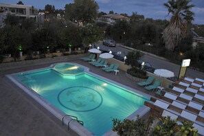 A swimming pool with a geometric design, surrounded by lounge chairs and umbrellas, with a road and buildings in the background. - Makis Louvlis Luxury Apartments (Corfu)