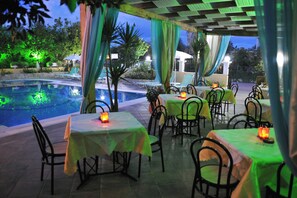 An outdoor dining area with tables and chairs, a pool, and greenery in the background. - Makis Louvlis Luxury Apartments (Corfu)
