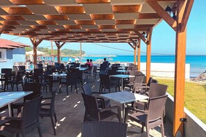An outdoor patio with tables and chairs, overlooking a beach and ocean. - Angela Beach Hotel (Corfu)