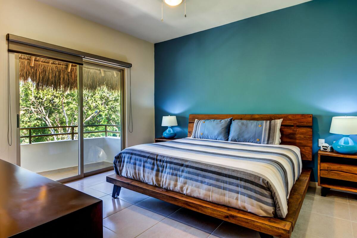 A bedroom with a large bed, two bedside tables with lamps, a wooden bench, and a sliding glass door leading to a balcony.