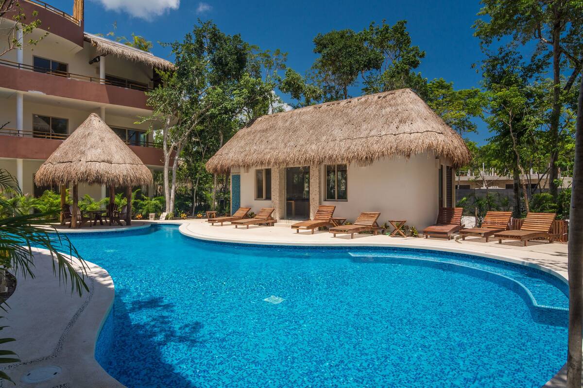 A resort pool with two thatched-roof huts, lounge chairs, and a clear blue pool surrounded by tropical vegetation.