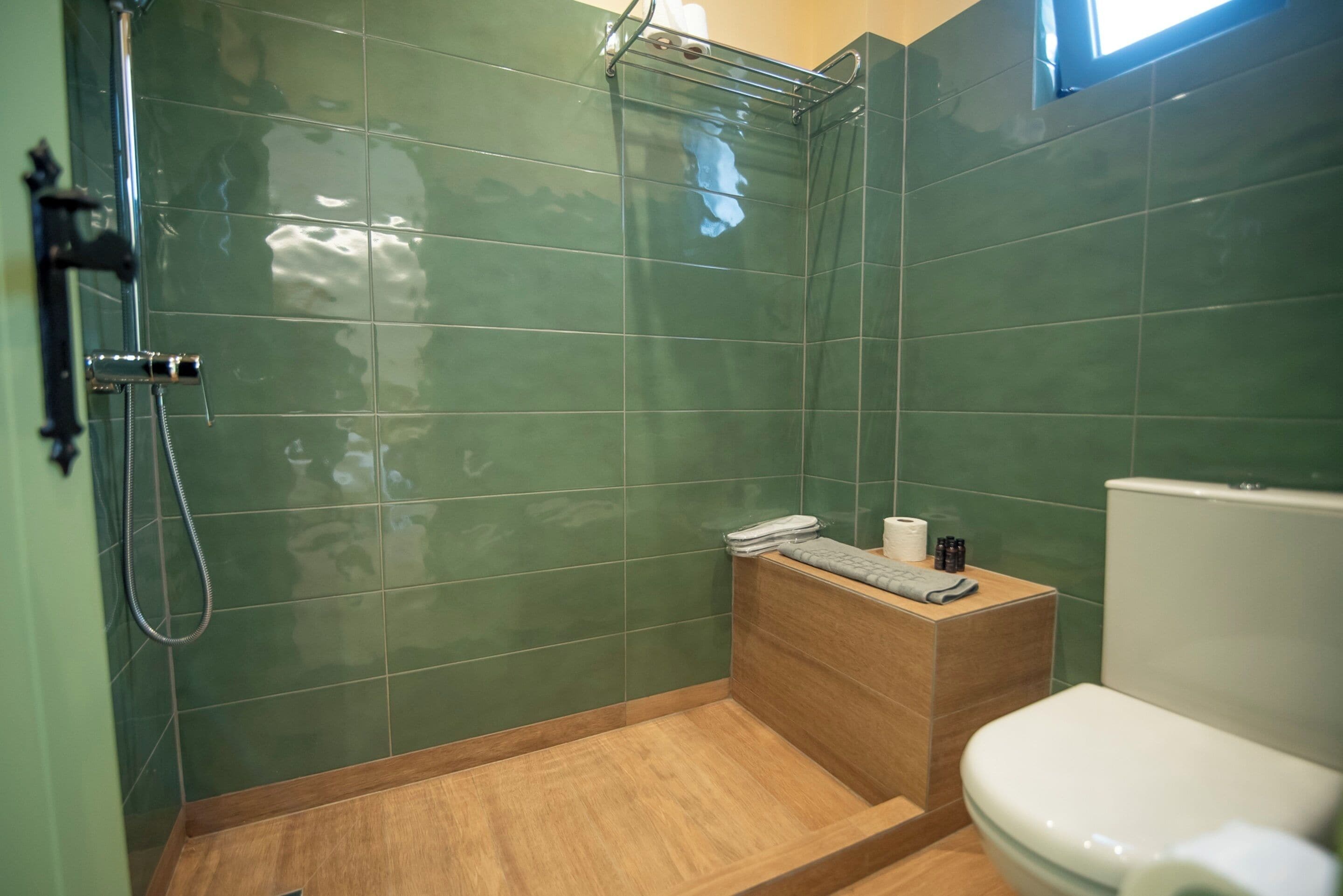 A bathroom with green tiles, a wooden shower area, a toilet, and a small shelf with toiletries.