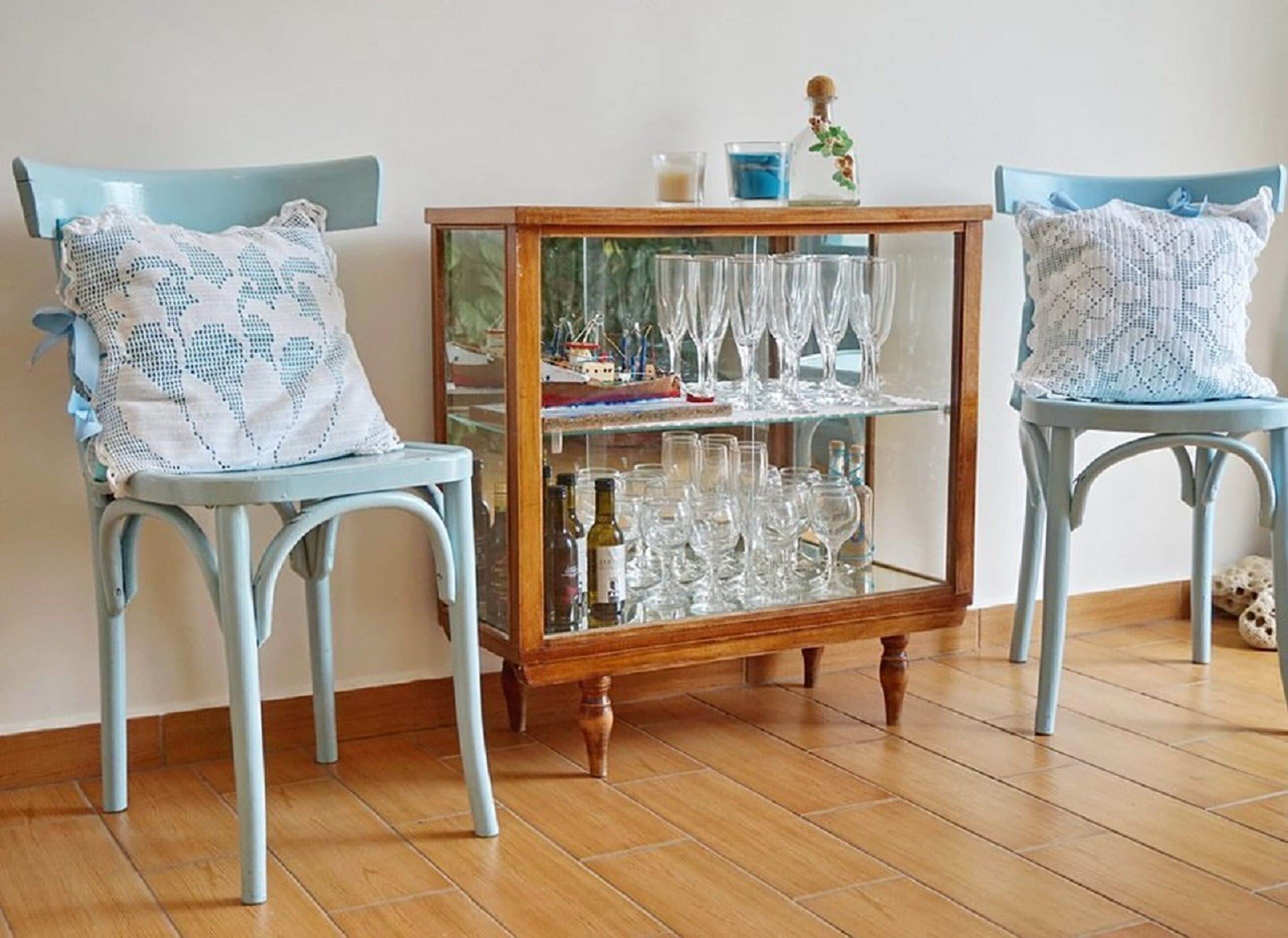 Two light blue chairs with white patterned cushions, a wooden cabinet with glass doors displaying wine glasses and bottles, and a small blue candle on top.