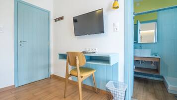 Una habitación con piso de madera, una puerta azul, un escritorio con una silla, un televisor de pantalla plana, una lámpara amarilla y un baño con espejo y lavamanos.