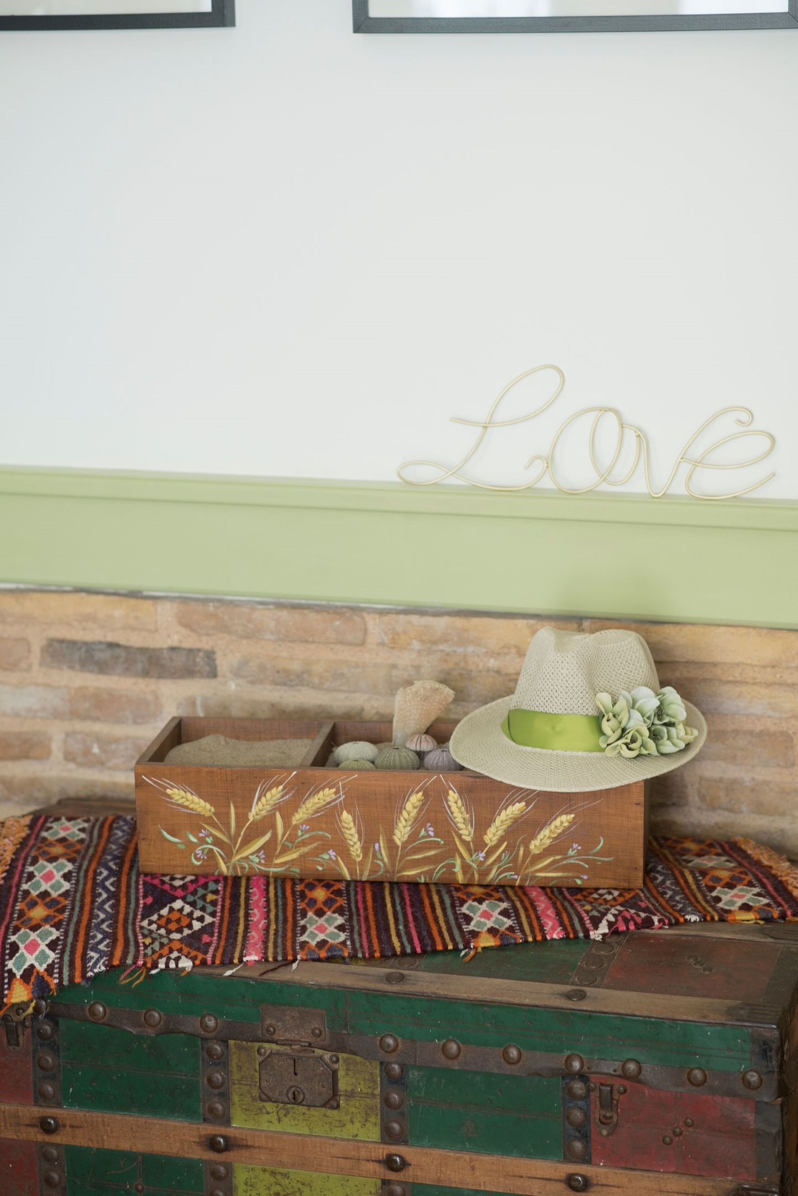 Un cofre de madera rústica con un sombrero tejido y objetos decorativos encima, frente a un muro de ladrillo con un letrero que dice 'Love'.