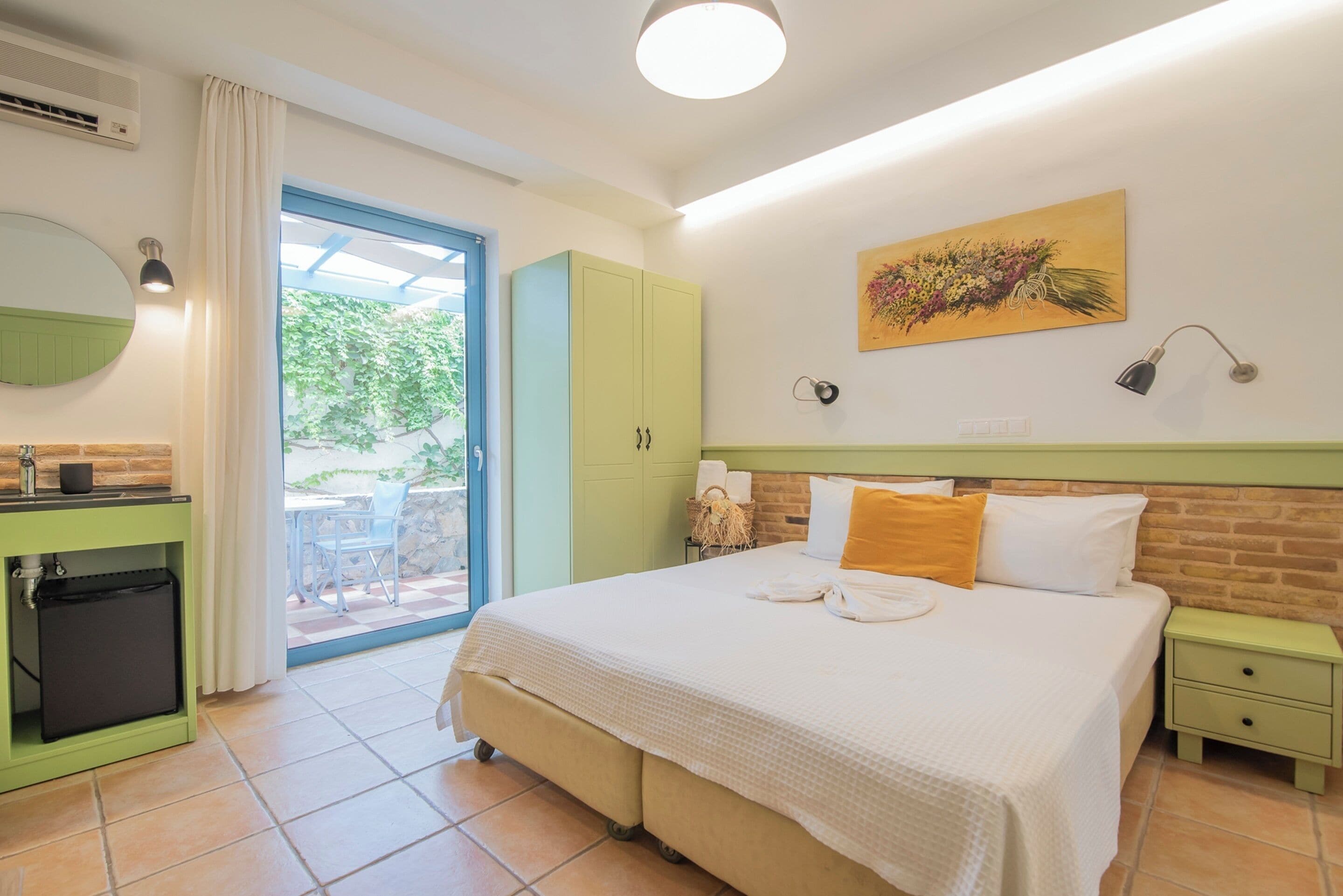 A bedroom with a bed, a green wardrobe, a small table with a lamp, and a window with a view of greenery.