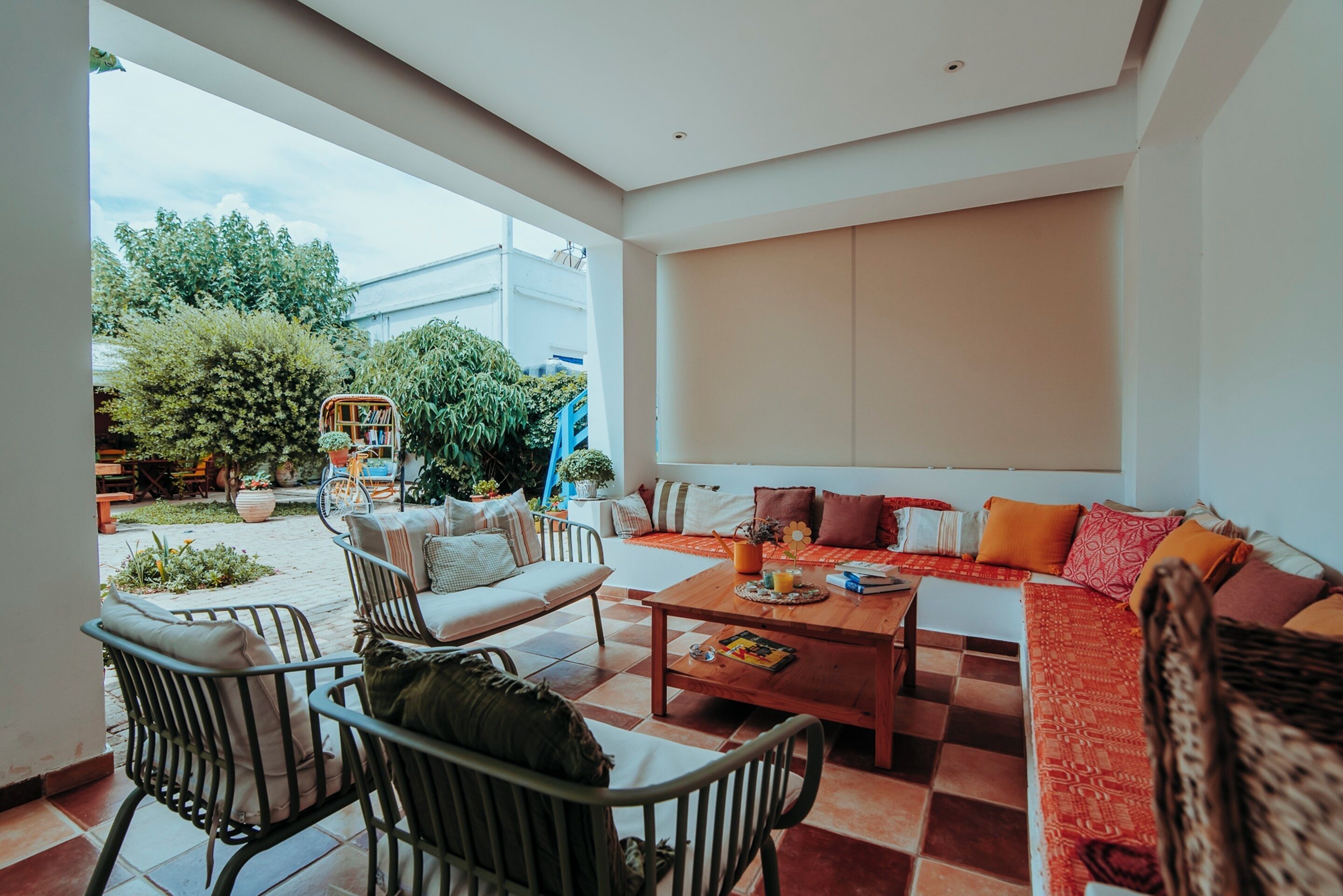 A spacious living area with a sofa set, a coffee table, and a dining table. There are chairs around the tables, and a view of the outdoors is visible through large windows.