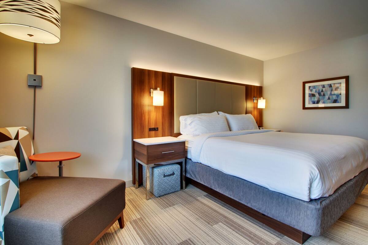 A hotel room with a large bed, a bedside table, a chair, a lamp, and a wall-mounted artwork.