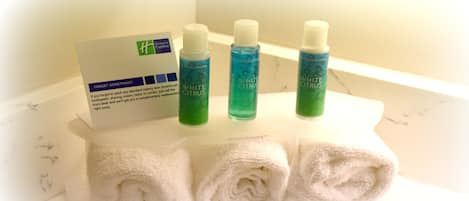 A bathroom with three bottles of white citrus shampoo, a white towel, and a card offering complimentary toiletries.