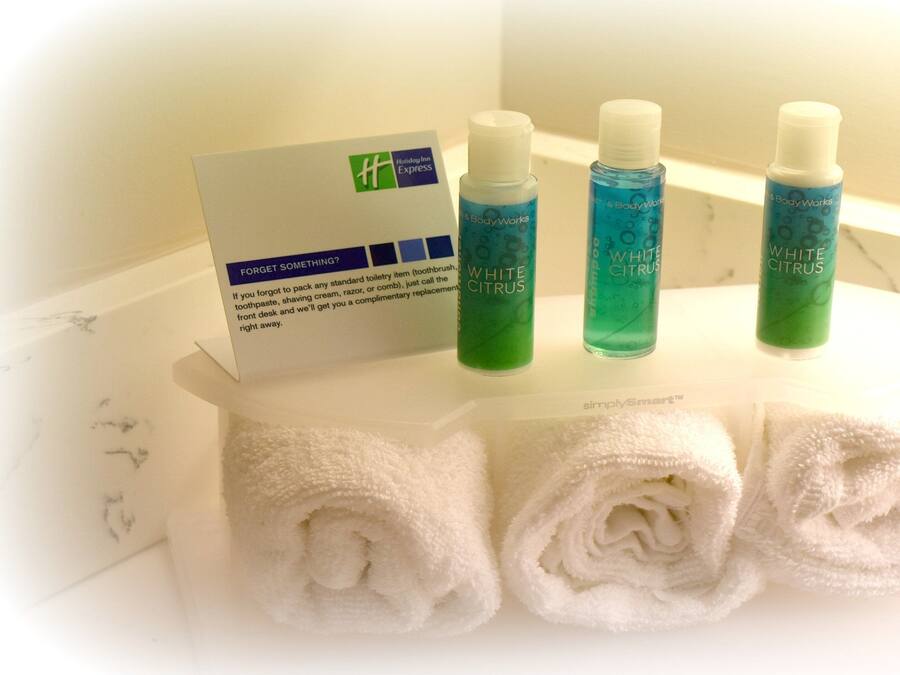 A bathroom with three toiletries on a shelf, a complimentary toiletries card, and two rolled towels.