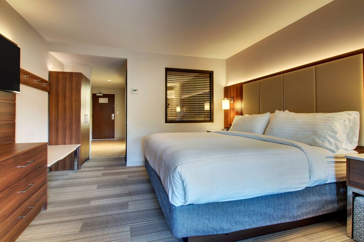 A modern hotel room with a large bed, a wooden dresser, a flat-screen TV, and a wall-mounted light fixture.
