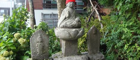 A stone statue of a seated figure with a red hat, surrounded by greenery and two stone markers.