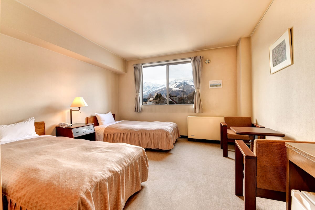 Habitación de hotel con dos camas, un escritorio, una silla, una lámpara, una ventana con vista a las montañas y un cuadro en la pared.