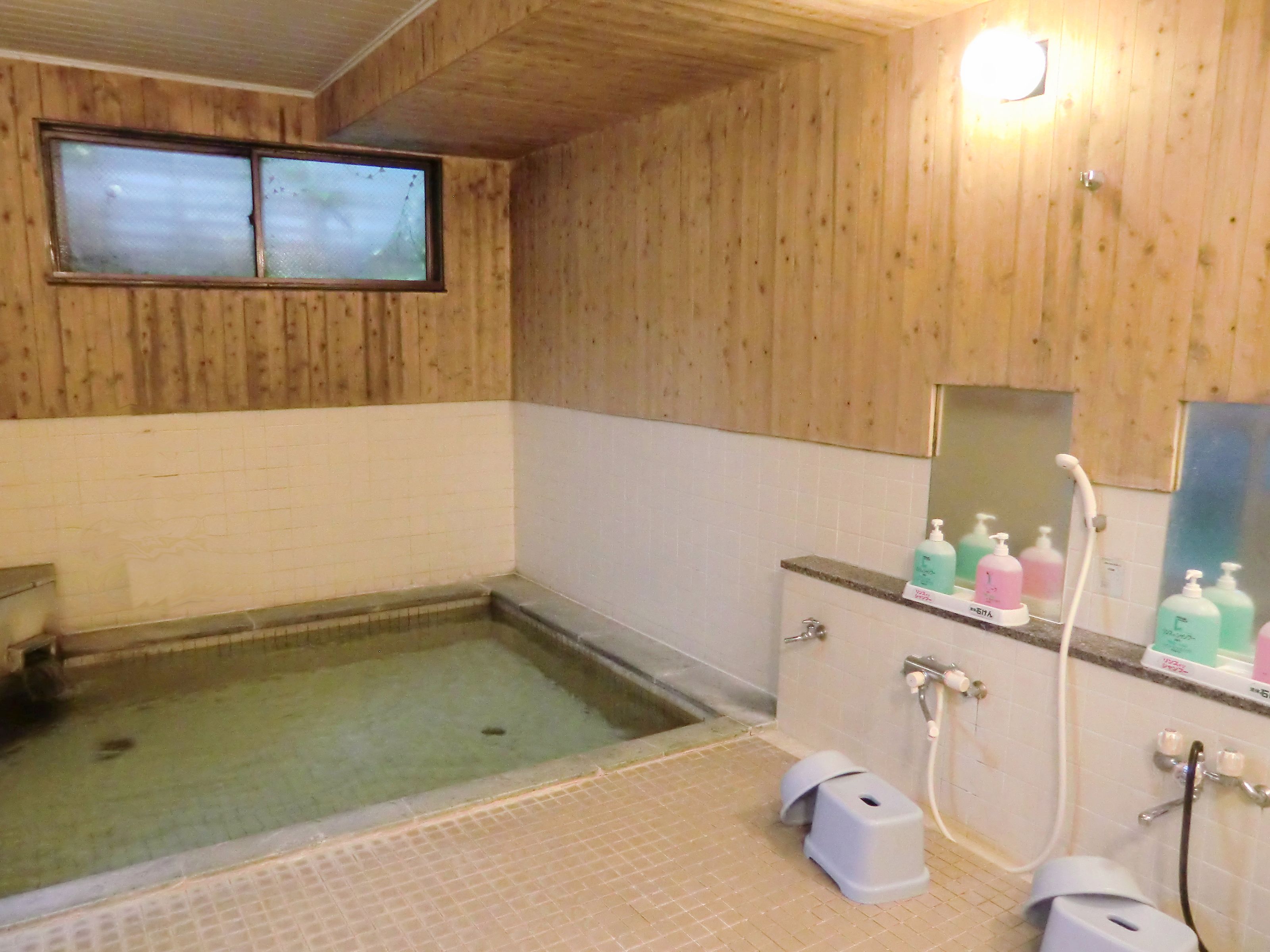A wooden-paneled bathroom with a small window, a tiled floor, and a shower area with a control panel and soap dispensers.