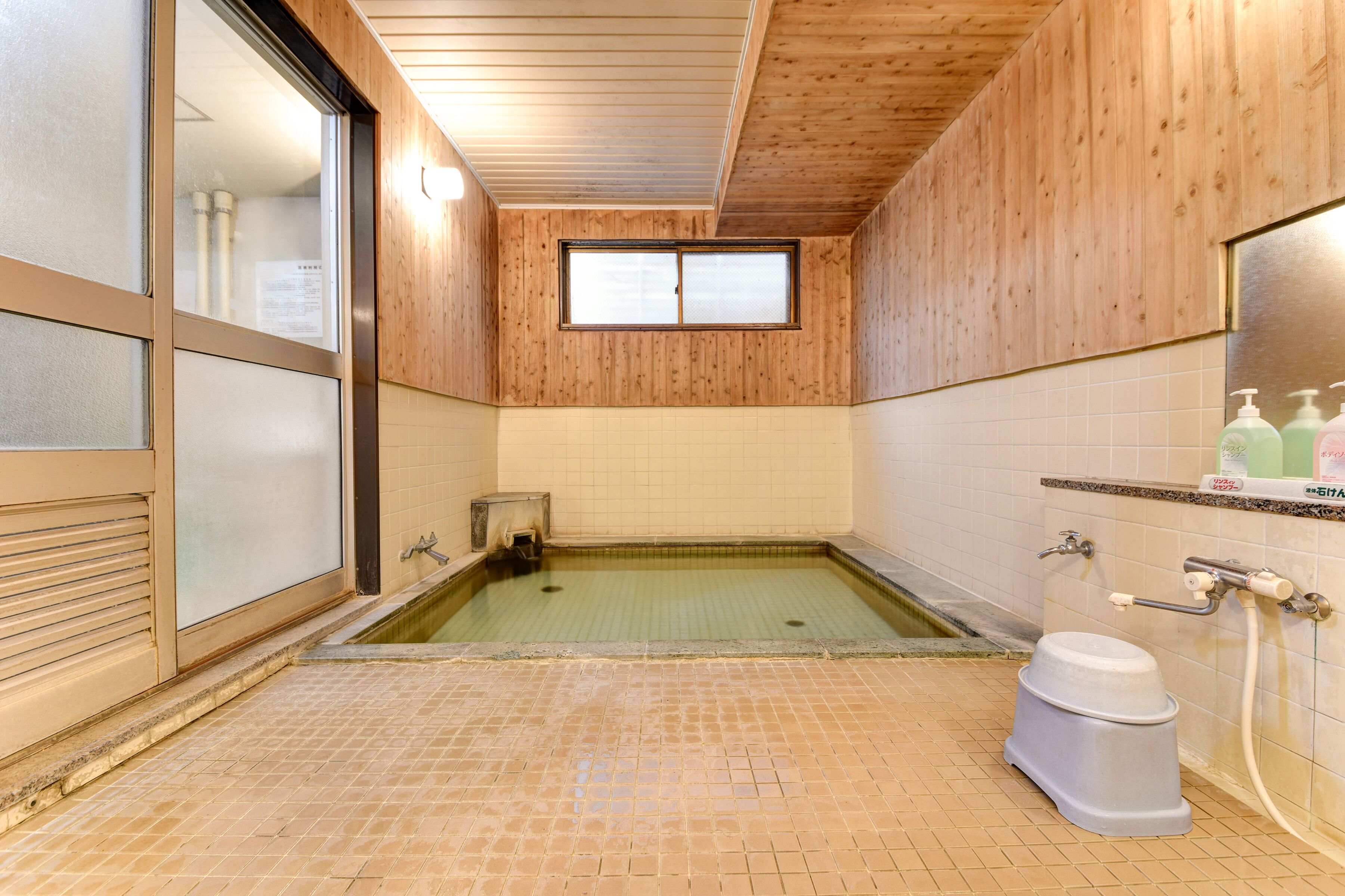 A small, tiled bathroom with a wooden ceiling, a window, a toilet, and a sink with soap dispensers.