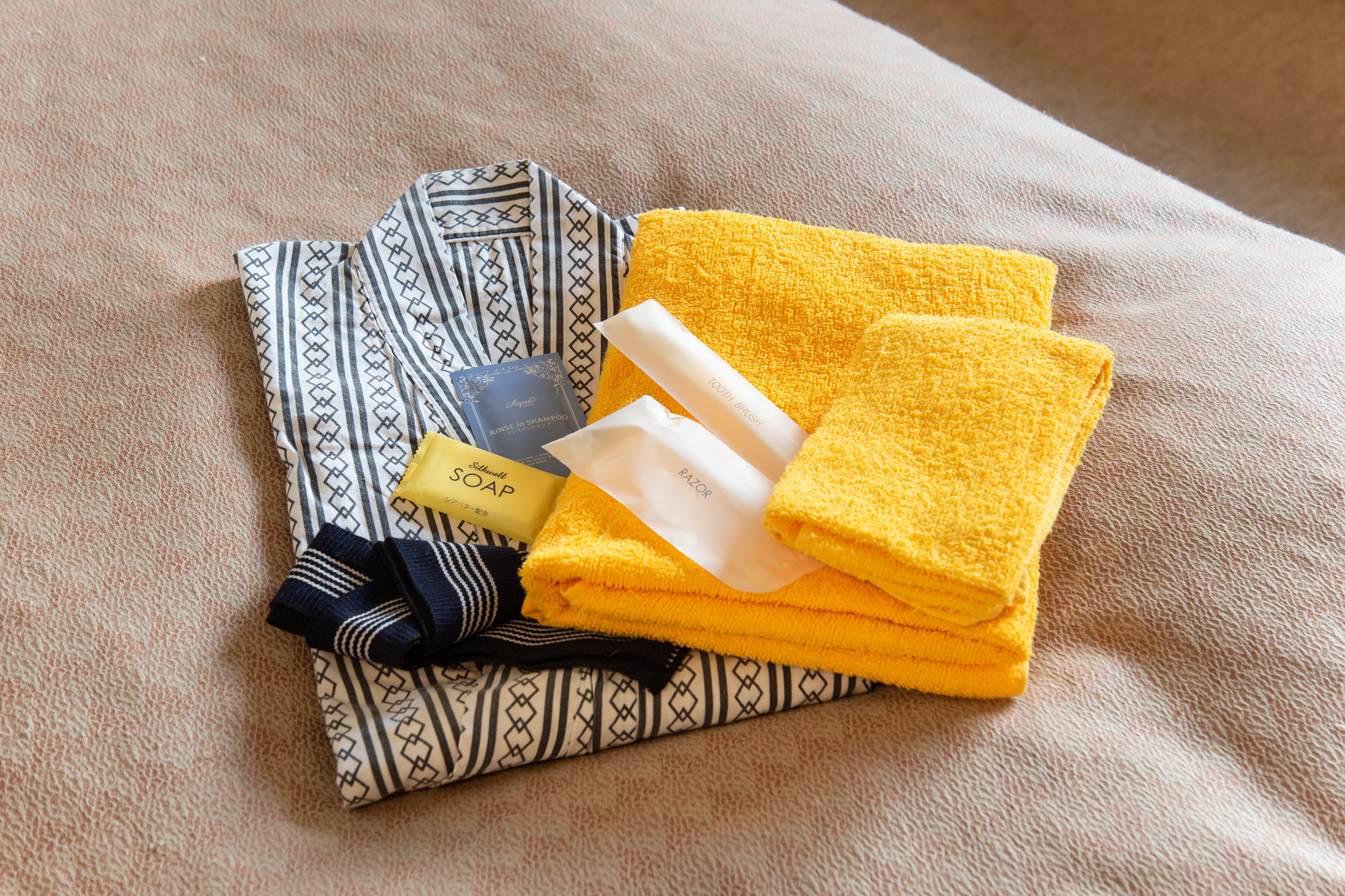 A neatly folded towel, a bar of soap, a bottle of shampoo, and a folded piece of clothing on a textured surface.