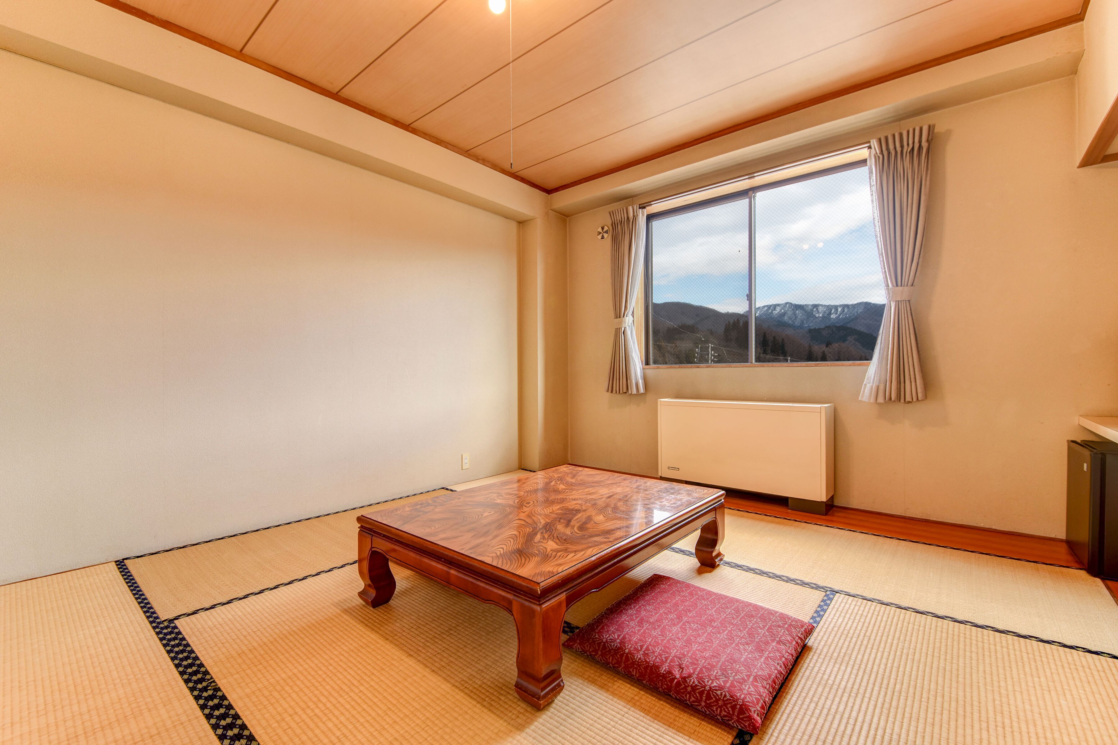 Een kamer met tatami vloer, een houten tafel, een rode kussen, een raam met gordijnen en een radiator.
