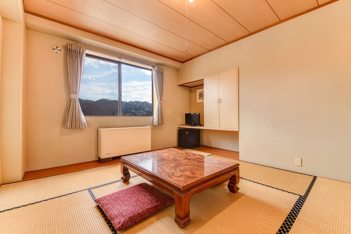 Una habitación con suelo de tatami, una mesa de madera, un radiador, una ventana con cortinas, un televisor y un armario.