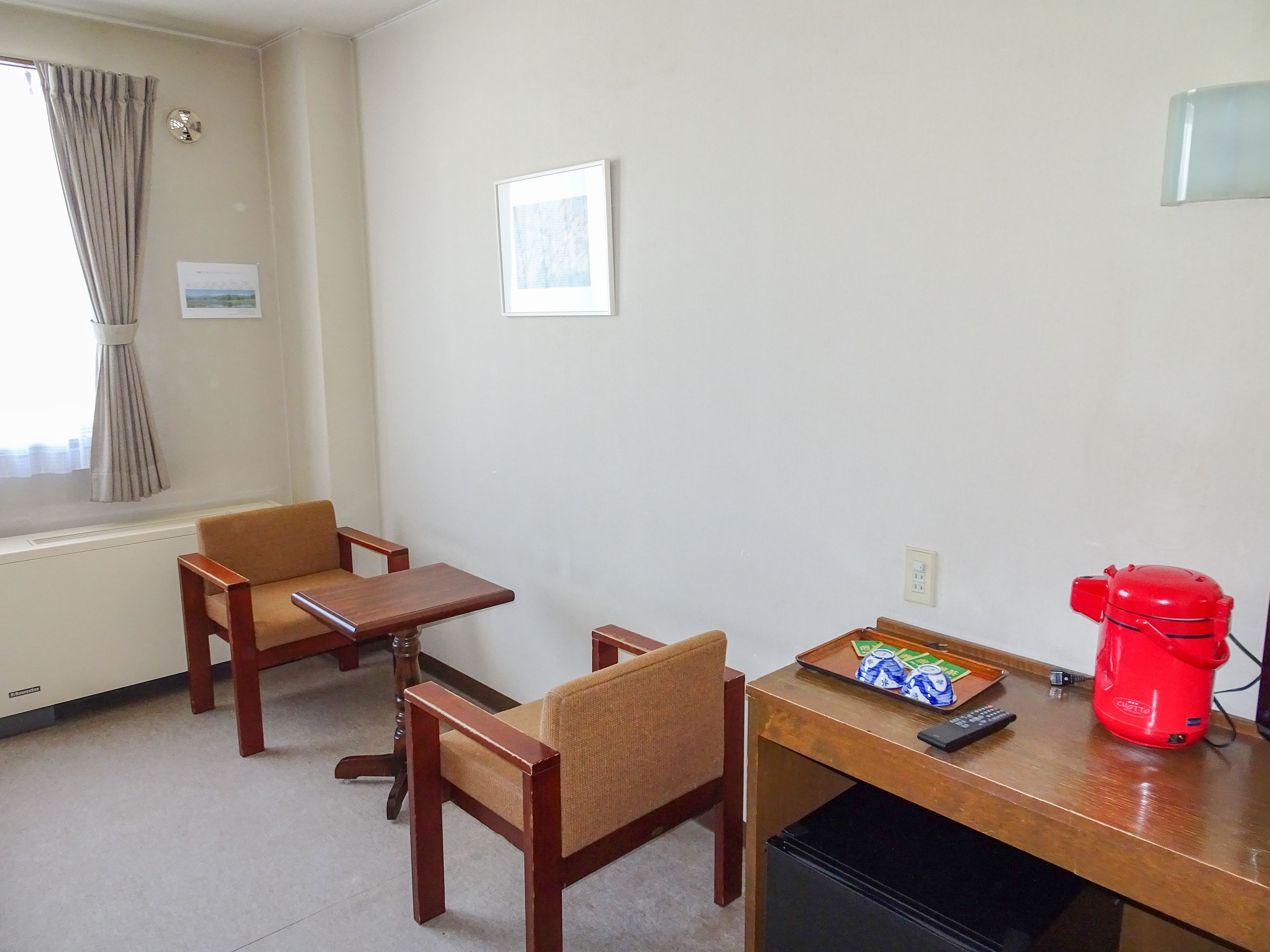 A hotel room with two wooden chairs, a small table, a red kettle, and a refrigerator.