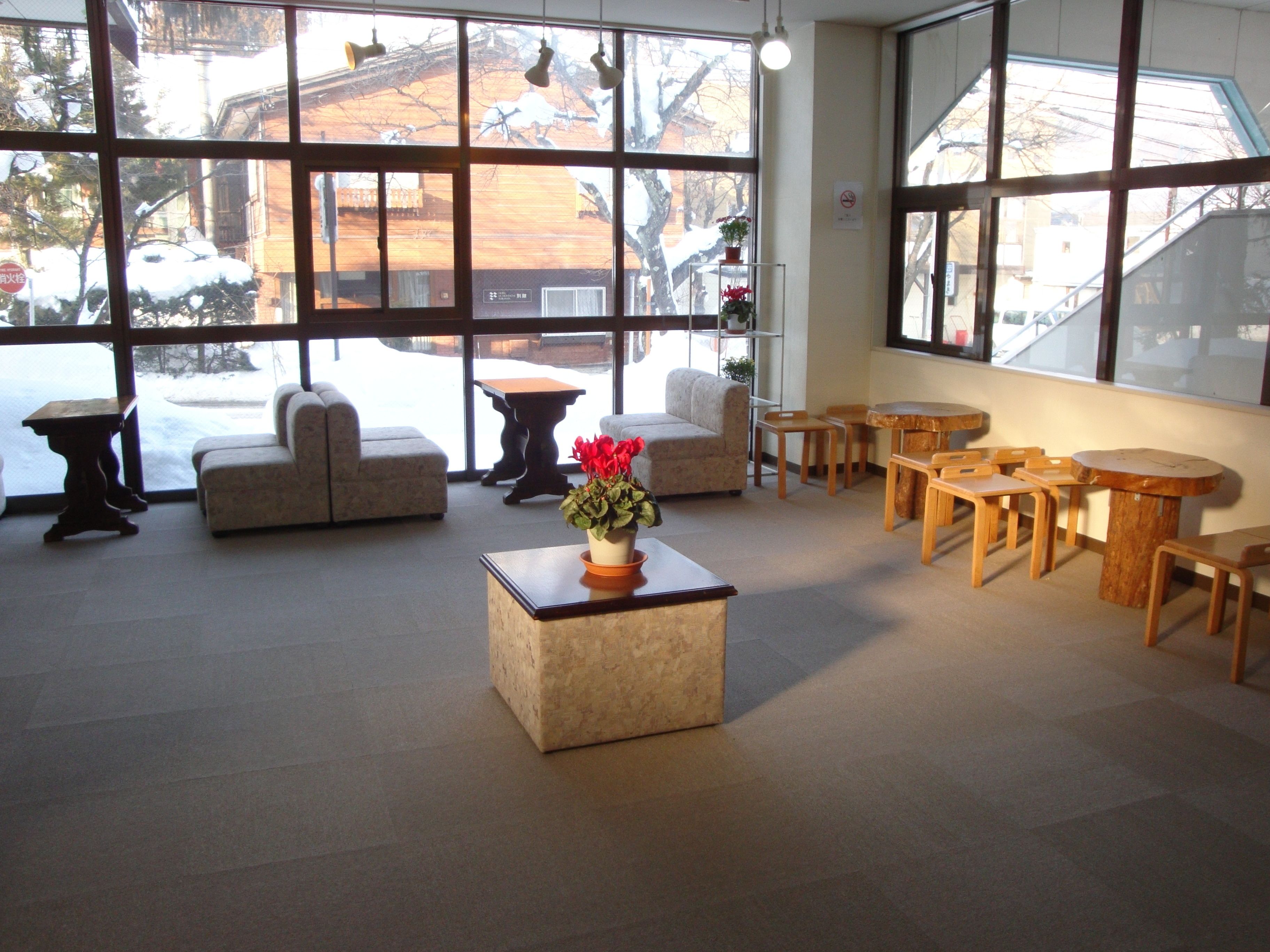 Un hall d’entrée spacieux, doté de grandes fenêtres, d’une table basse centrale agrémentée d’un arrangement floral, et de plusieurs espaces de détente comprenant des canapés et des tables en bois.