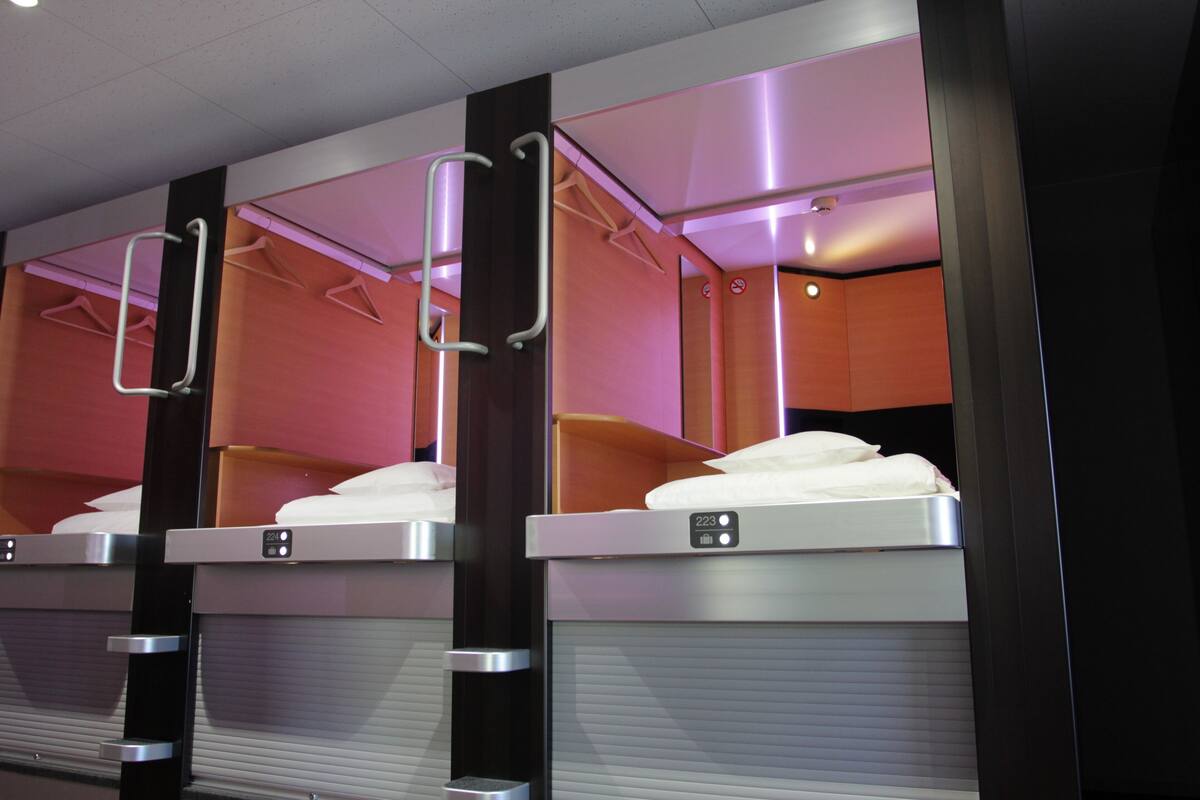 Une rangée de lits superposés avec des barrières métalliques, chaque lit est équipé d’un panneau de commande, et la pièce présente un design moderne avec un éclairage rose.