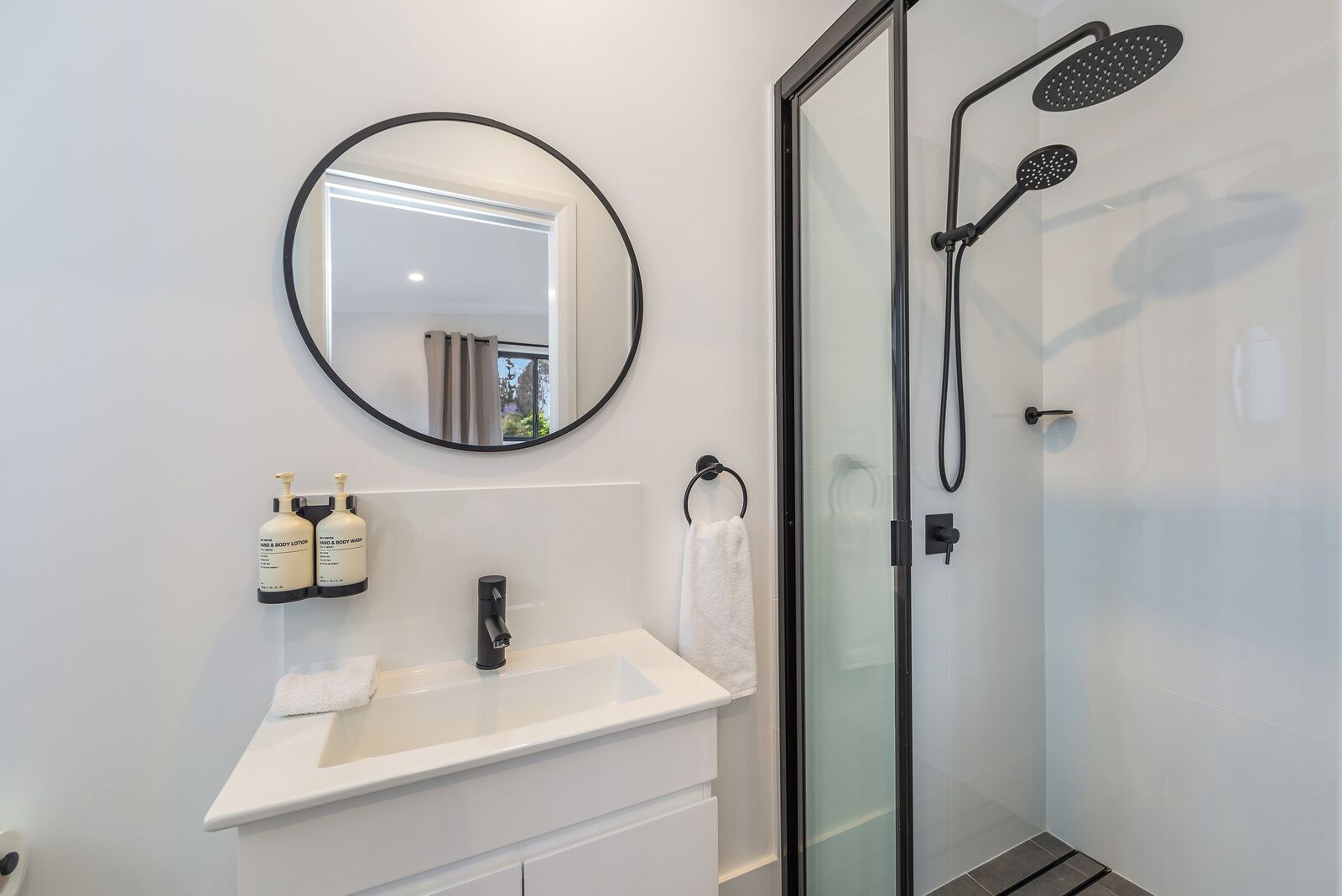 Een moderne badkamer met een ronde spiegel, een witte wasbak, twee flessen hand- en douchezeep, een handdoekhouder en een douchecabine met glazen wanden.