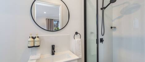 A modern bathroom with a round mirror, white sink, two bottles of hand and body wash, a towel ring, and a glass-enclosed shower.