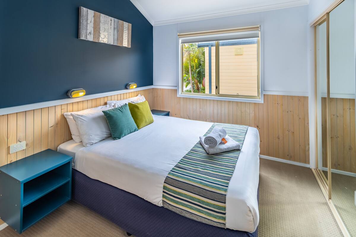 A bedroom with a bed, a nightstand, a window with a view, and a wooden wall panel.