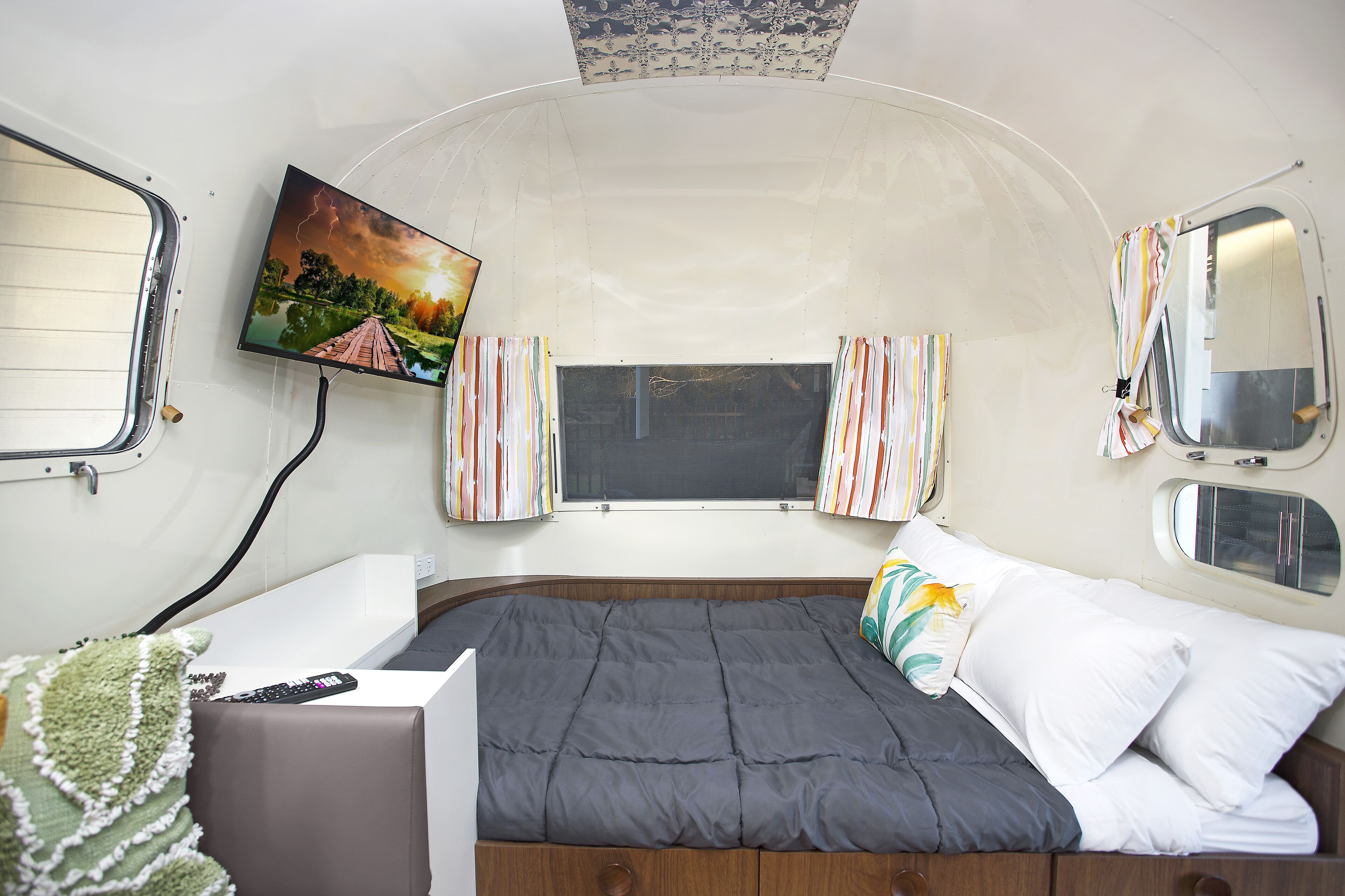 A compact RV bedroom with a bed, a TV mounted on the wall, and a window with curtains.
