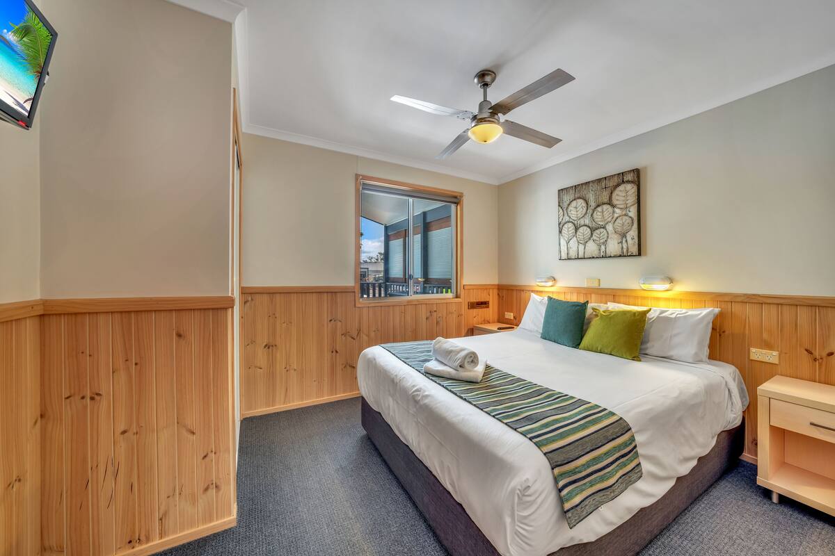 A hotel room with a bed, a ceiling fan, a nightstand, a mirror, and a window with blinds.
