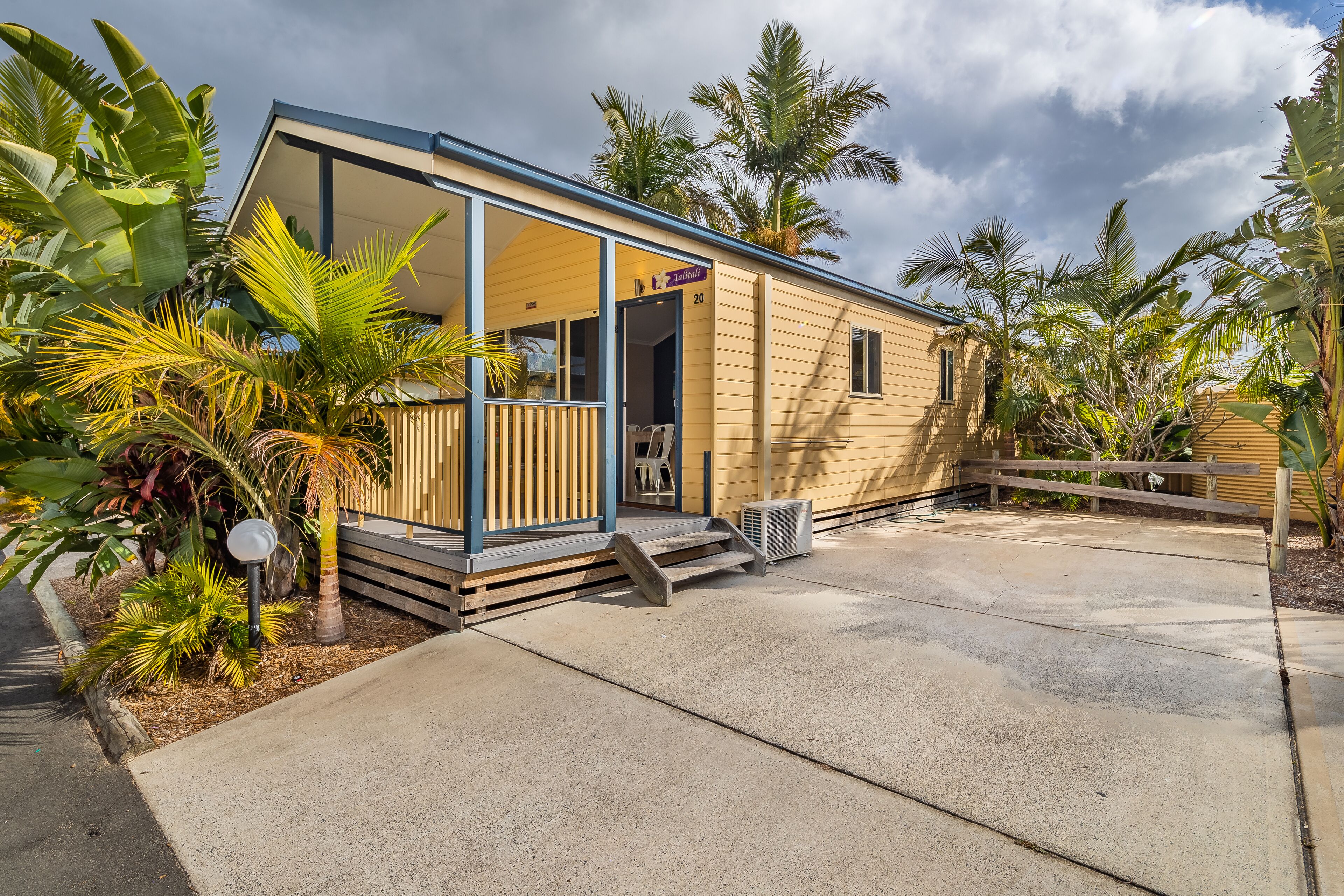 A single-story, yellow building with a glass door entrance, surrounded by tropical plants.